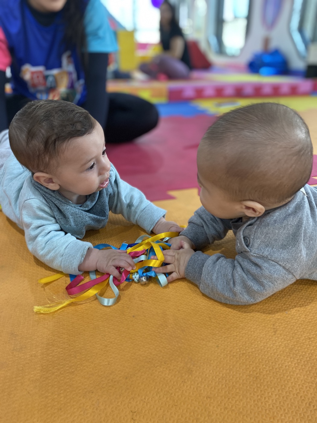 Dois bebês deitados no tatame explorando fitas coloridas juntos durante uma aula da Baby Gym Itaim Bibi